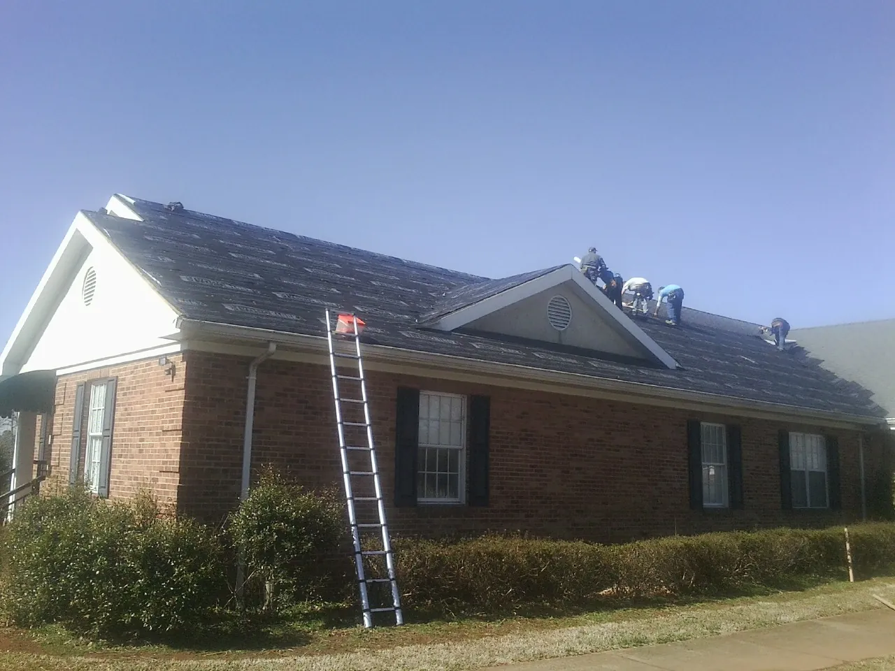 Crew tearing shingles off a brick ranch roof with a ladder set — Metal Roof Repair work by our crew in Martinsville